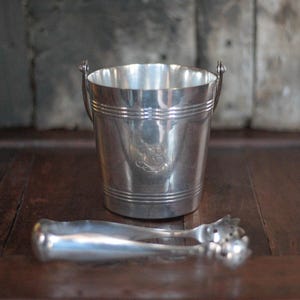 May include: A silver ice bucket with a handle and a pair of ice tongs. The bucket has a decorative emblem on the front and horizontal bands around the body. The tongs are also silver and have decorative ends.