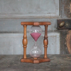 May include: An hourglass with a wooden frame and clear glass bulbs. The sand inside is a deep pink color. The hourglass is set on a dark surface against a weathered wooden door.