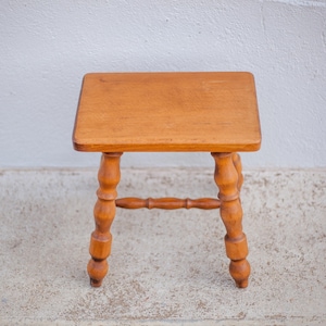 May include: A small wooden stool with a square top and turned legs. The stool is made of light brown wood and has a simple design.