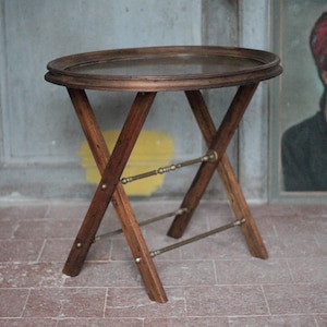 May include: A vintage, oval-shaped wooden side table with a glass top. The table has a folding, X-shaped leg design with brass accents. The wood has a warm, brown tone. The table is approximately 24 inches in diameter and 20 inches tall.