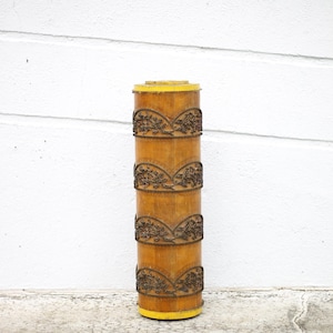 May include: A tall, cylindrical bamboo container with decorative floral bands. The container is primarily light brown with yellow accents on the top and bottom. The bands are a darker brown, adding contrast to the design.