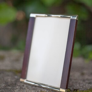 May include: A rectangular picture frame with a dark wood border and a gold-colored metal trim. The frame is angled slightly, displaying a white interior. The background is blurred, with green foliage and a stone surface.