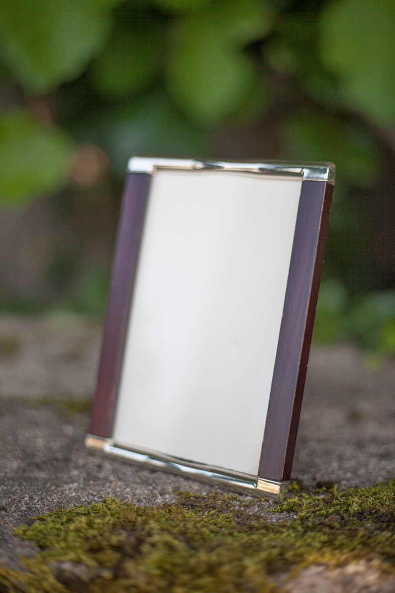 May include: A rectangular picture frame with a dark wood border and a gold-colored metal trim. The frame is angled slightly, displaying a white interior. The background is blurred, with green foliage and a stone surface.
