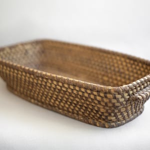May include: A rectangular woven basket made of natural brown and light brown reeds. The basket has two handles on the sides.