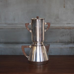Peut inclure: Une cafetière vintage en argent avec des poignées en bois et un bouton en bois sur le dessus. La cafetière a une conception en deux parties, avec une base conique et une section supérieure cylindrique. Les poignées sont de couleur marron foncé.