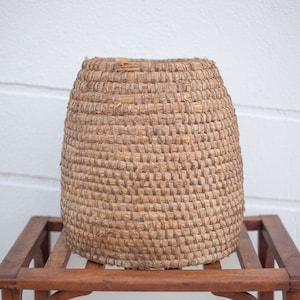 May include: A large, round, woven basket made of natural materials. The basket is brown and has a natural, rustic look. It is sitting on a wooden table.