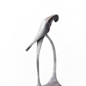 Op de afbeelding: Een zilveren metalen sculptuur van een vogel die op een standaard zit. De vogel zit in een pose alsof hij naar beneden kijkt.