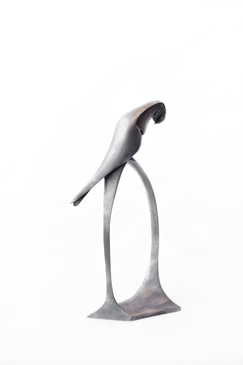 Op de afbeelding: Een zilveren metalen sculptuur van een vogel die op een standaard zit. De vogel zit in een pose alsof hij naar beneden kijkt.