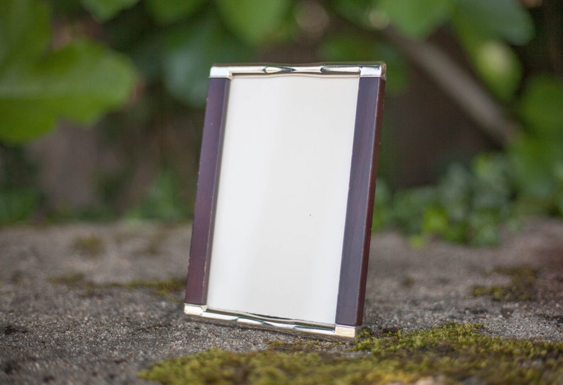 May include: A rectangular photo frame with a dark wood finish and silver-colored metal trim. The frame is empty, with a white interior, and is standing upright on a stone surface. The background is blurred with green foliage.