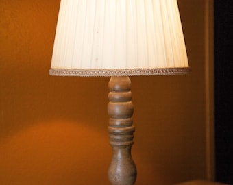 May include: A wooden table lamp with a white pleated lampshade and a brown wooden base. The lamp is turned on and casting a warm glow.