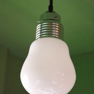 May include: A white light bulb with a silver metal base hangs from a black cord against a green background.
