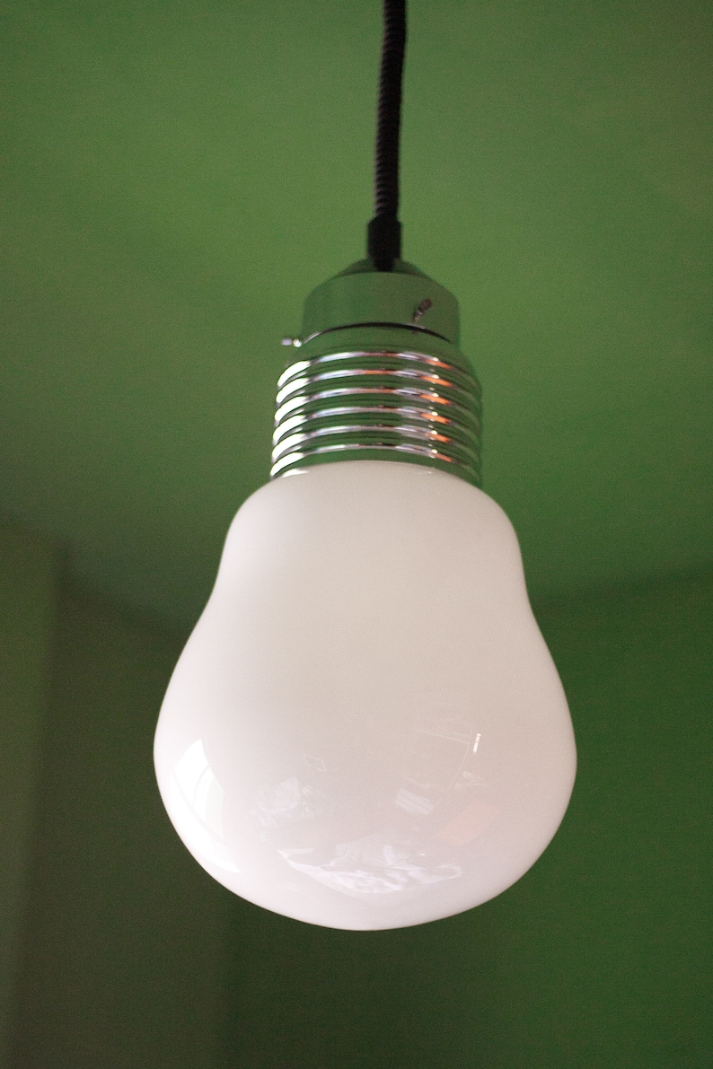 May include: A white light bulb with a silver metal base hangs from a black cord against a green background.