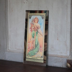Peut inclure: Impression encadrée rectangulaire représentant une femme en robe verte fluide, tenant des fleurs. L'œuvre d'art a un style vintage avec une palette de couleurs sourdes de verts, de roses et de crèmes. Le cadre est en métal foncé simple. L'impression est appuyée contre une surface en bois vieilli.