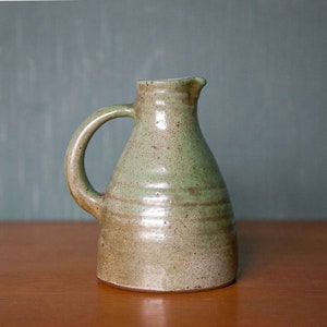 Könnte beinhalten: Ein kleiner, grüner und brauner Keramik Krug mit Henkel. Der Krug hat eine gesprenkelte Glasur und eine leicht abgerundete Form.
