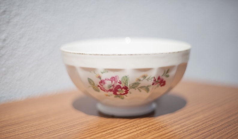 K&ouml;nnte beinhalten: Eine wei&szlig;e Keramiksch&uuml;ssel mit einem Blumendesign mit rosa und roten Blumen. Die Sch&uuml;ssel hat einen geriffelten Rand und ein glattes, wei&szlig;es Innere.