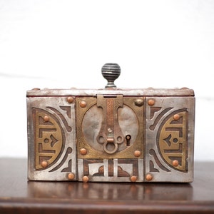 May include: A rectangular metal box with intricate details. The box features a silver-colored body with brass accents, including decorative patterns and rivets. A keyhole and ornate latch are centered on the front, and a ridged knob sits atop the lid.