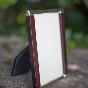 May include: A rectangular picture frame with a dark wood border and silver-colored metal trim. The frame has a white interior and a black easel back. The frame is standing on a stone surface with a blurred green background.