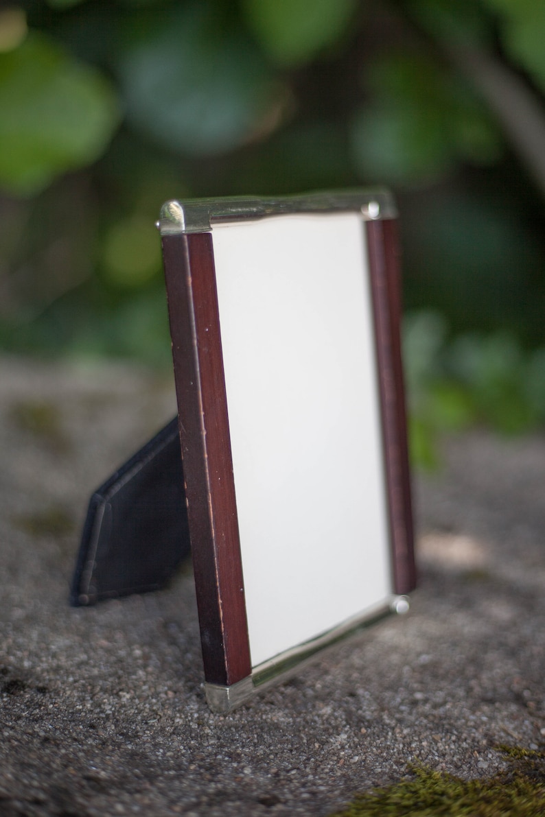 May include: A rectangular picture frame with a dark wood border and silver-colored metal trim. The frame has a white interior and a black easel back. The frame is standing on a stone surface with a blurred green background.