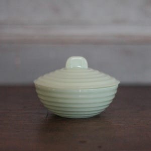 Op de afbeelding: Een kleine, lichtgroene keramische container met een deksel. De container en het deksel hebben een geribbeld, concentrisch ontwerp. Het deksel heeft een kleine, vierkante handgreep. De container staat op een donkerbruin oppervlak.
