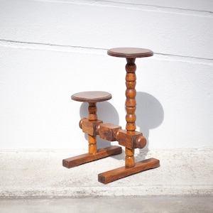 Peut inclure: Un support de plante en bois avec deux étagères rondes. Le support a un design unique avec une série de boutons en bois tournés sur les pieds et un support en forme de croix entre les étagères.