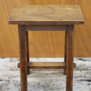 Può includere: Uno sgabello di legno semplice con un piano rettangolare e quattro gambe. Lo sgabello è in legno marrone scuro e ha un aspetto rustico.