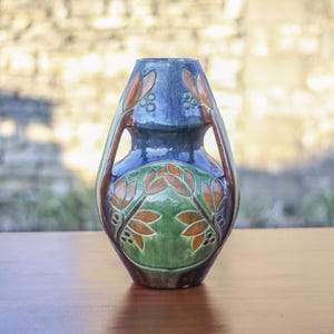 Könnte beinhalten: Eine blaue und grüne Keramikvase mit floralem Muster. Die Vase hat zwei Henkel und einen runden Korpus.
