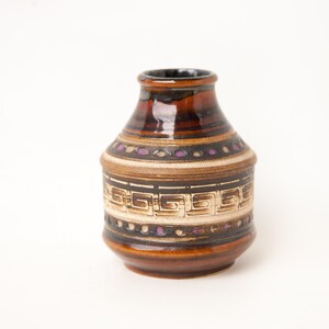 Peut inclure: Un petit vase en céramique brun avec un motif géométrique en blanc et brun. Le vase a un corps arrondi et un col étroit.