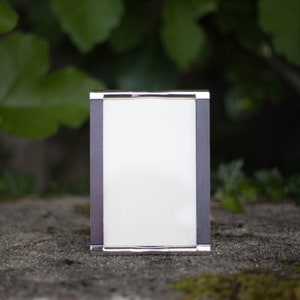 May include: A rectangular silver-framed photo frame with a dark gray border and a white interior. The frame is sitting on a stone surface with green foliage in the background.
