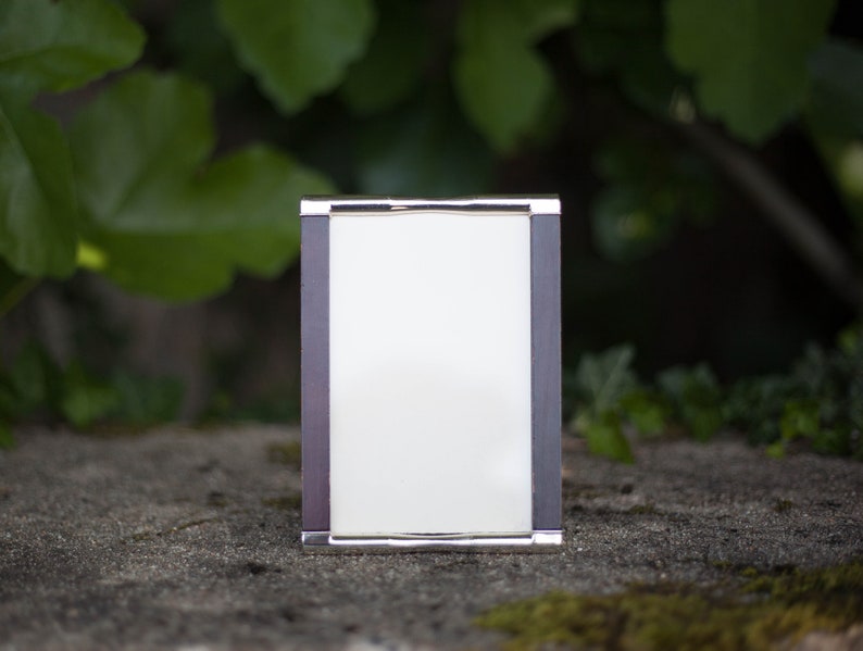 May include: A rectangular silver-framed photo frame with a dark gray border and a white interior. The frame is sitting on a stone surface with green foliage in the background.