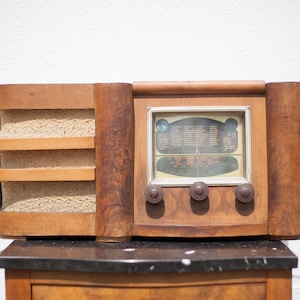Op de afbeelding: Een vintage houten radio met een luidsprekerrooster en een wijzerplaat met stationsnamen. De radio heeft drie knoppen en staat op een zwart marmeren oppervlak.
