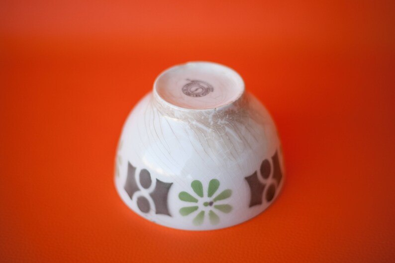 May include: White ceramic bowl with a brown, green, and white geometric design. The bowl has a slightly textured surface and a small brown stamp on the bottom.
