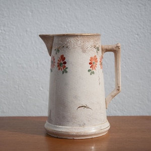 May include: A white ceramic pitcher with a floral design. The pitcher has a handle and a wide mouth. The flowers are red, orange, and green.