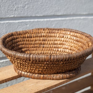 May include: A large, round, woven basket made of natural materials. The basket is brown and has a slightly flared rim. It is sitting on a wooden surface.