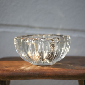 Puede incluir: Un cuenco de vidrio transparente con un diseño acanalado se encuentra sobre una superficie de madera. El cuenco está vacío y tiene un ligero tono dorado.