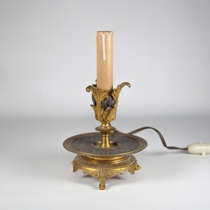 Peut inclure: Une lampe de table dorée avec une ampoule en forme de bougie et une base décorative. La base a un dessus rond et orné et trois pieds.