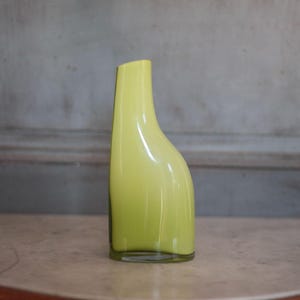 May include: A tall, slender, lime-green glass vase with a curved, organic shape. The vase has a slightly angled opening and a clear glass base. The smooth, glossy surface reflects light, highlighting its unique form. This decorative vase is suitable for floral arrangements.