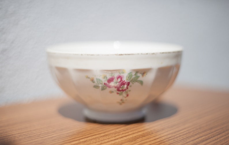 K&ouml;nnte beinhalten: Eine wei&szlig;e Keramiksch&uuml;ssel mit einem Blumendesign mit rosa und roten Blumen. Die Sch&uuml;ssel hat einen geriffelten Rand und steht auf einer Holzoberfl&auml;che.