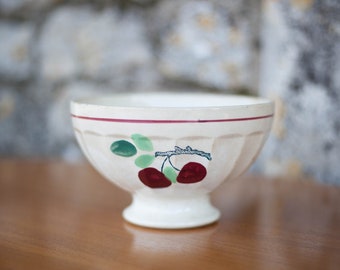 May include: A white ceramic bowl with a red rim and a cherry design. The bowl has a fluted edge and a pedestal base.
