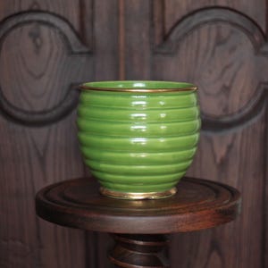 Peut inclure: Un pot de fleurs en céramique verte avec des rainures horizontales et une bordure dorée repose sur un piédestal en bois foncé. Le pot a une forme ronde et une finition brillante. L'arrière-plan est une porte en bois foncé.