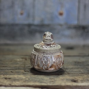 Puede incluir: Un pequeño tarro de cerámica hecho a mano con tapa. El tarro es beige con detalles marrones y un diseño tallado y texturizado. La tapa presenta una pequeña cara estilizada. El tarro está sobre una superficie de madera envejecida.