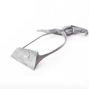 Op de afbeelding: Een zilverkleurig metalen gereedschap met een gebogen handvat en een plat, rechthoekig blad. Het gereedschap is ontworpen voor gebruik in de leerbewerking.