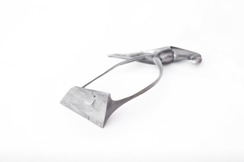 Op de afbeelding: Een zilverkleurig metalen gereedschap met een gebogen handvat en een plat, rechthoekig blad. Het gereedschap is ontworpen voor gebruik in de leerbewerking.