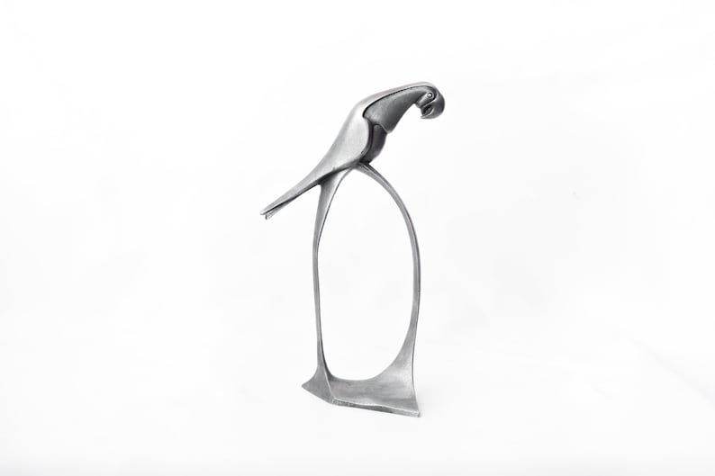 Op de afbeelding: Een zilveren metalen sculptuur van een papegaai die op een gebogen basis staat. De kop van de papegaai is naar de zijkant gedraaid en zijn snavel is gesloten.