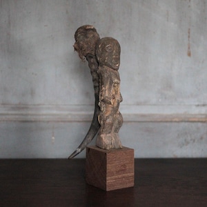 May include: A weathered, wooden sculpture of two figures, one atop the other, mounted on a small, square wooden base. The sculpture has a rough, textured surface and a natural, brown color. The background is a muted gray wall.