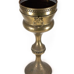Peut inclure: Un grand pot à fleurs en laiton antique avec une finition martelée et des détails complexes. Le pot a un haut rond et large avec un bord décoratif et une seule poignée en anneau. La base est un pied rond et large.