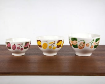 May include: Three white ceramic bowls with different fruit designs. The smallest bowl has a pink and brown cherry design, the middle bowl has a yellow and brown lemon design, and the largest bowl has a green and brown peach design.