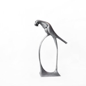 Op de afbeelding: Een zilveren metalen sculptuur van een vogel die op een gebogen standaard zit. De vogel kijkt naar beneden met zijn kop ingetrokken. Het beeld heeft een modern, minimalistisch ontwerp.