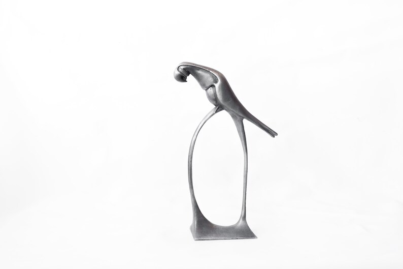 Op de afbeelding: Een zilveren metalen sculptuur van een vogel die op een gebogen standaard zit. De vogel kijkt naar beneden met zijn kop ingetrokken. Het beeld heeft een modern, minimalistisch ontwerp.