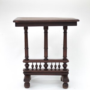Peut inclure: Petite table en bois brun foncé avec un plateau rectangulaire et des détails ornés. La table comporte trois supports verticaux et une rampe inférieure décorative avec de petits accents arrondis. Les pieds sont également arrondis.