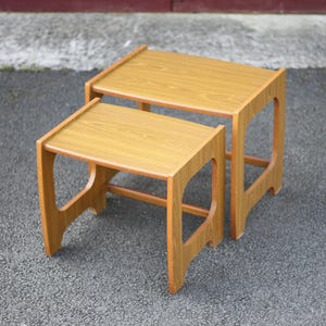 Puede incluir: Un conjunto de dos mesas nido de madera con un acabado marrón claro. Las mesas tienen un diseño sencillo y moderno de mediados de siglo con una tapa rectangular y un marco abierto único.
